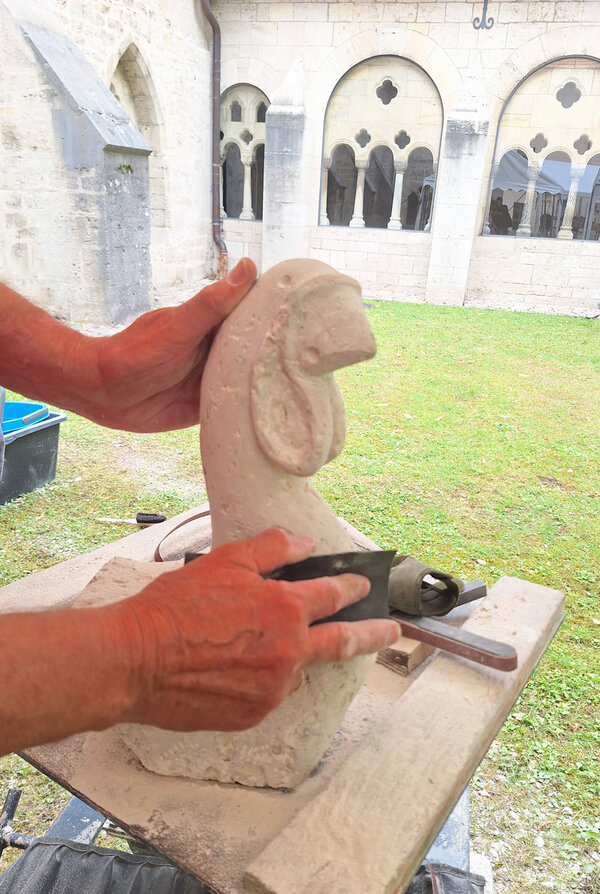 Bild vergr&ouml;&szlig;ern: Das Bild zeigt die Hände eines Steinmetzes, der eine Statue aus Stein schnitzt. Im Hintergrund sind gotische Fenster eines Klosters zu sehen.